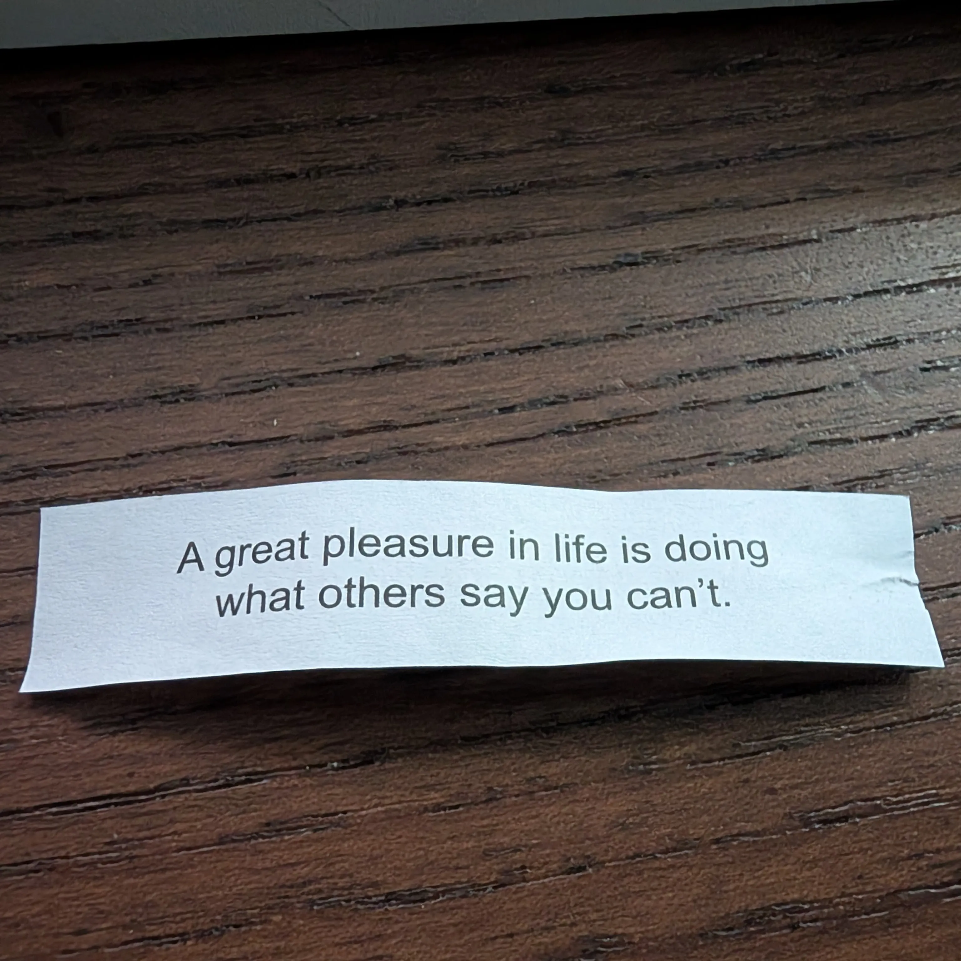 A piece of paper on a wooden desk. The paper reads "A great pleasure in life is doing what others say you can't.